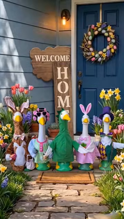 Five white decorative goose statues lined up on a front porch, dressed in various spring and Easter-themed costumes including bunny suits and festive hats. They stand on a stone path surrounded by colorful spring flowers, a tall wooden 'Welcome Home' sign, and a blue door adorned with an Easter egg wreath.