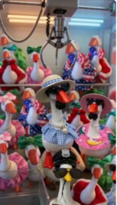 Inside a claw machine game, a crowd of white toy geese dressed in assorted colorful outfits and accessories, including sunglasses, hats, and dresses. A metal claw hangs suspended directly above them.