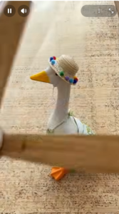 Side profile of a white toy goose wearing a small straw hat decorated with colorful pom-poms